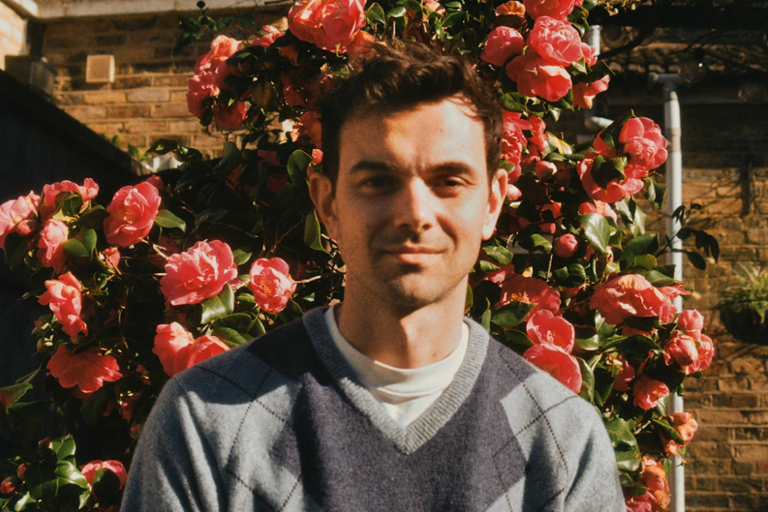 Portrait of Tom Iansek of #1 Dads wearing a blue argyle sweater, posing outdoors in front of vibrant pink camellia flowers in bloom.